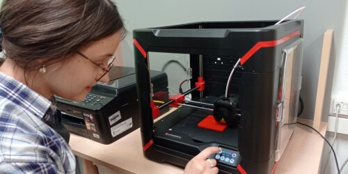 Uczennica zmienia parametry na wyświetlaczu drukarki 3D. Drukarka drukuje czerwony prostokąt.