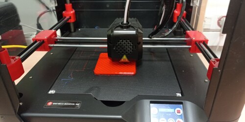 Czarna drukarka 3D drukuje czerwony sześcian.