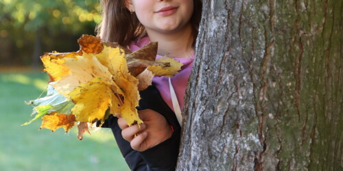 Dziewczynka schowana za pniem drzewa trzyma liście w dłoni