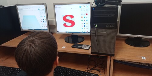 Uczeń modyfikuje parametry wydruku 3D litery S. W tle ekran komputera z wyświetloną czerwoną literą S,