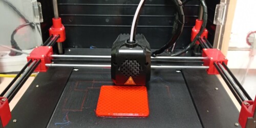 Czarna drukarka 3D drukuje czerwony sześcian.