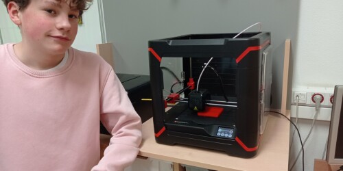 Uczeń stoi przy drukarce 3D. Drukarka 3D drukuje czerwony prostokąt.