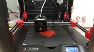 Drukarka 3D drukuje czerwone serce.W tle czarna obudowa drukarki.