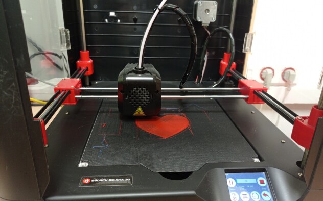 Drukarka 3D drukuje czerwone serce.W tle czarna obudowa drukarki.