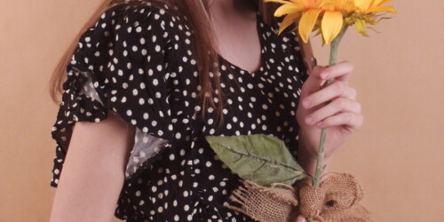 Uczennica w ciemnej sukiencie pozuje do zdjęcia z kwiatem. Na drugim planie tło.