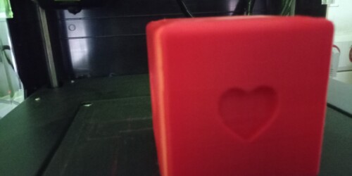 Sześcienny czerwony klocek wykonany z PLA. Serce na przedniej ścianie. W tle drukarka 3D.