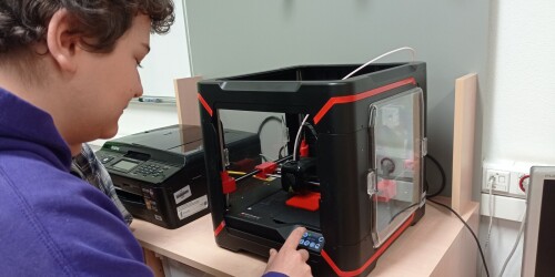 Uczeń w fioletowej bluzie zmienia na wyświetlaczu drukarki 3D parametry wydruku. W tle drukarka 3D drukuje czerwony sześcian.