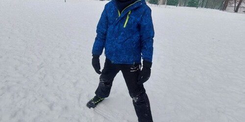 Dziecko w rozkroku, w ciepłym ubraniu, stoi na boisku pokrytym śniegiem.