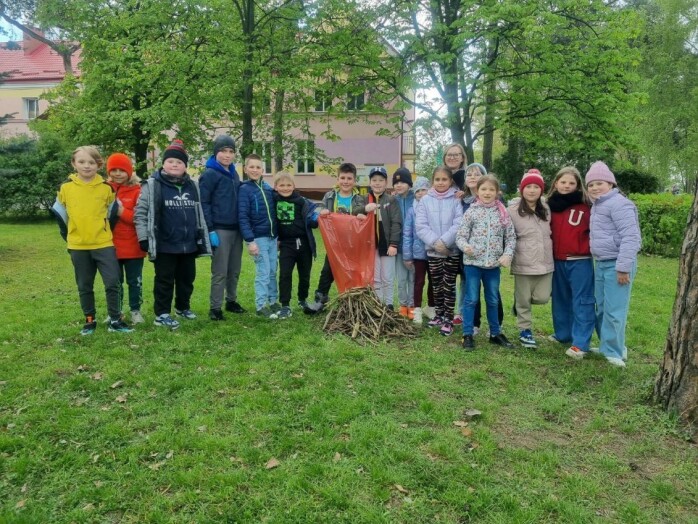 Uczniowie klasy IIB pozują w parku szkolnym podczas akcji Sprzątanie Świata ułozony z patyków kopiec. Jeden uczeń trzyma czerwony worek na śmieci. W tle drzewa i budynek szkoły.
