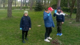 Trzech uczniów klasy IIIb w parku szkolnym podczas akcji Sprzątania Świata poszukują gałązek oraz śmieci. W tle drzewa pokryte zielonymi liśćmi.