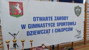 Baner informacyjny, niebieski napis na białym tle. Napis na banerze: otwarte zawody w gimnastyce sportowej dziewcząt i chłopców.
