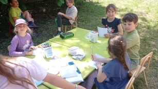 Uczniowie siedzą przy stolikach w zielonym ogrodzie szkolnym. Przygotowują prace plastyczne. W tle zielone krzewy i drzewa.