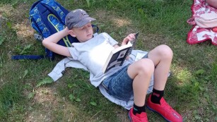 Uczeń leży na trawie oparty o plecak i czyta książkę.